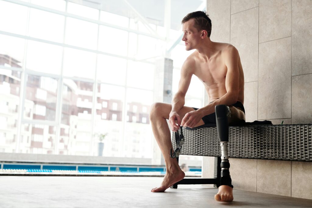 Young man with prosthetic leg sitting by a swimming pool, preparing for a swim indoors.